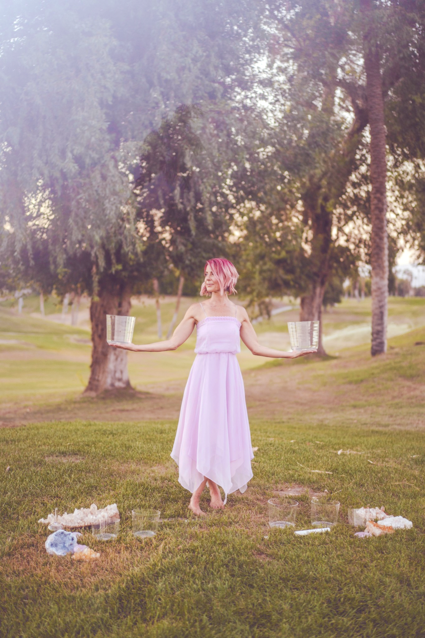 Bri standing barefoot in a lavender dress in soft sunlight, holding a crystal bowl in each outstretched hand, with more bowls and crystals on the grass.