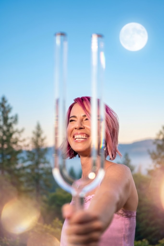 Bri smiling outdoors and holding a tuning fork toward the camera, with the moon above her.